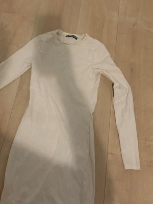 Beige långärmad kortklänning - En enkel och stilren beige kortklänning med lång ärm och rund halsringning. Klänningen har en kroppsnära passform och är tillverkad i ett mjukt bomullsmaterial. Perfekt för dig som gillar minimalistisk och clean stil.