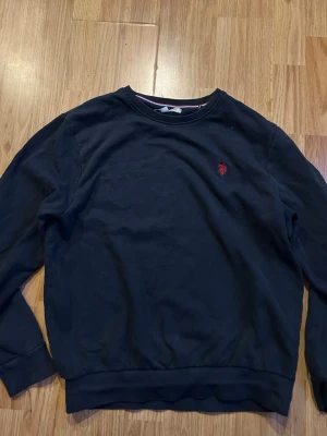 Mörkblå sweatshirt från U.S. Polo Assn. - Klassisk mörkblå sweatshirt från U.S. Polo Assn. med rund halsringning och röd broderad logga på bröstet. Tröjan har långa ärmar och ribbade muddar vid ärmslut och nederkant. Perfekt för en avslappnad och stilren look.