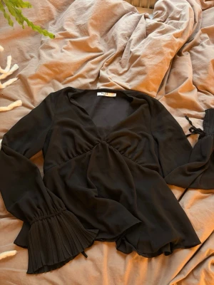 Svart omlottblus från NA-KD - Stilren svart omlottblus från NA-KD med långa, vida ärmar. Blusen har en djup v-ringning och knytband i midjan. Köpt för 450. Super fin! Skriv för fler bilder eller frågor!🥰