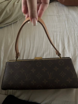 Liten handväska - Säljer en liten handväska i klassiskt brunt mönster. Väskan har en avlång, boxig form med beige läderhandtag och guldfärgade metalldetaljer. Några repor på sidorna se bild 4-5! Därav priset. 