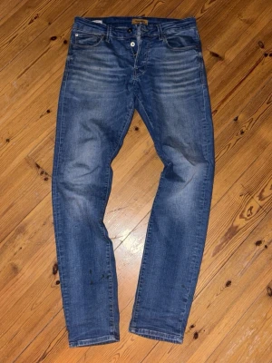 Blå slim fit jeans från Jack n Jones  - Säljer ett par klassiska blå slim fit jeans från Jack n Jones.  Jeansen snygga slitningar framtill för en avslappnad look. Tillverkade i mjuk denim med stretch för extra komfort. Perfekta till vardagsstilen!                   En liten liten slitning vid baksida lår, se på andra bilden! Annars inga defekter 