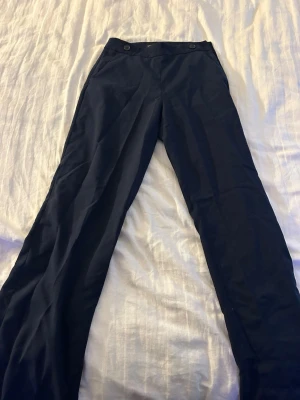 Mörkblå kostymbyxor med raka ben - Snygga mörkblå kostymbyxor med raka ben och hög midja. Byxorna har två knappar framtill och en diskret ögla bak i linningen. Klassisk och stilren design, perfekta till skjorta eller kavaj.