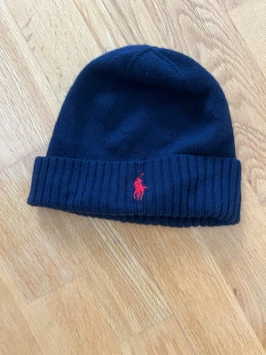 Mörkblå mössa från Polo Ralph Lauren - Snygg mörkblå mössa från Polo Ralph Lauren i mjuk merinoull. Klassisk ribbad kant och röd broderad logga framtill. Perfekt accessoar för kalla dagar och enkel att matcha med olika stilar.