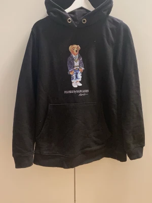 Svart Hoodie Polo Bear - Svart hoodie från Ralph Lauren med Polo Bear-tryck på bröstet. Klassisk känguruficka framtill, huva med dragsko och ribbade muddar. Materialet känns mjukt och bekvämt, perfekt för en avslappnad stil. Ikonisk björn i kostym ger en unik touch. Storleken är S men skulle säga att den sitter mer som typ en M. Vid fler frågor kom privat💯😁