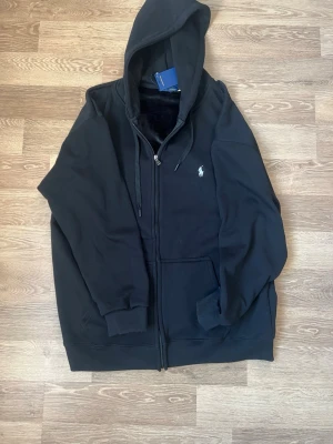 Svart hoodie från Polo Ralph Lauren - Svart hoodie med dragkedja från Polo Ralph Lauren. Klassisk design med huva och dragsnören, två fickor framtill och den ikoniska vita logotypen broderad på bröstet. Tillverkad i mjuk bomullsblandning som känns skön mot huden.