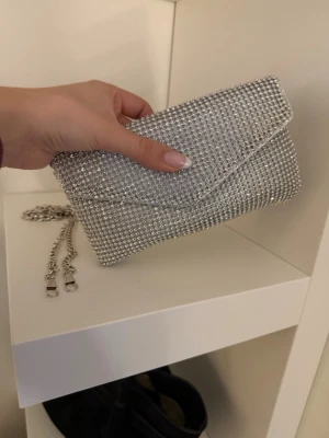 Silvrig kuvertväska med strass - Snygg silvrig kuvertväska täckt av glittrande strass och med en avtagbar kedjerem i metall. Väskan har en klassisk kuvertform och stängs med lock. Perfekt för att ge din outfit extra bling och stil.