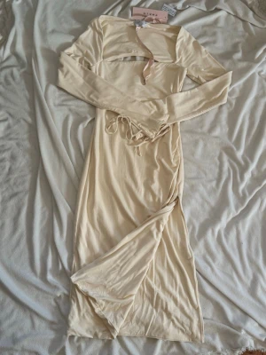 Långärmad beige klänning från Oh-polly - Trendig beige långklänning från Oh-polly med långa ärmar och snygga snörningsdetaljer vid handlederna. Klänningen har en unik cut-out upptill och är tillverkad i ett mjukt, stretchigt material som ger en smickrande passform.