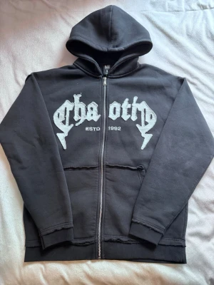 Svart hoodie med tryck från Black Squad sweterek z dzianiny - Svart hoodie från Black Squad med vit text och tryck på bröstet där det står 'chaoth' och 'ESTD 1992'. Hoodien har dragkedja framtill, två fickor och en klassisk huva. Relaxed fit och tillverkad i mjuk bomullsmix.Axelbredd: 60 Längd: 68 !!priset kan diskuteras!!