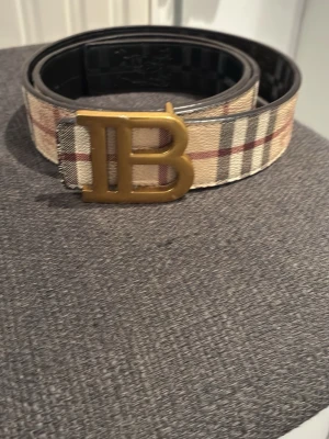 Rutig Burberry bälte med B-spänne - Snyggt bälte från Burberry med klassiskt rutigt mönster i beige, svart, rött och vitt. Bältet har ett stort guldfärgat B-spänne framtill som ger en lyxig känsla. Perfekt accessoar för att lyfta vilken outfit som helst. 110cm 