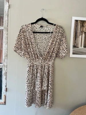 Beige kortklänning med leopardmönster - Söt kortklänning från GinaTricot i beige med svart leopardmönster, djup v-ringning och resår i midjan. Klänningen har korta ärmar och volangdetalj nedtill. Har inga tecken på användning mer än en liten lös söm vid bröstet (se sista bilden)🩷