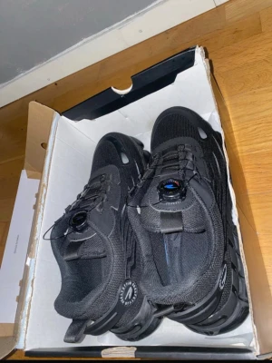 Svarta sneakers med snörningssystem - Säljer ett par svarta sneakers med robust sula och smart snörningssystem med snurrhjul. Skorna har mesh- och syntetmaterial för bra andningsförmåga och komfort. Perfekta för dig som vill ha en sportig och modern look till vardags eller träning.