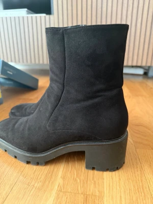 Svarta mockastövletter med klack - Snygga svarta stövletter i mocka med chunky sula och bred klack. Skorna har en minimalistisk design med rundad tå och dragkedja på insidan. Perfekta för dig som gillar en stilren look med lite edge.