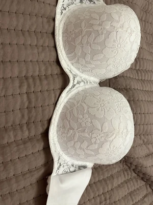Vit spets-bh med blommönster - Säljer en vit bh med vacker blommig spetsmönster. Bh:n har breda sidopartier som är gjort av osynlig plast och justerbara hakar i ryggen. Perfekt för dig som gillar romantiska detaljer och en feminin look.