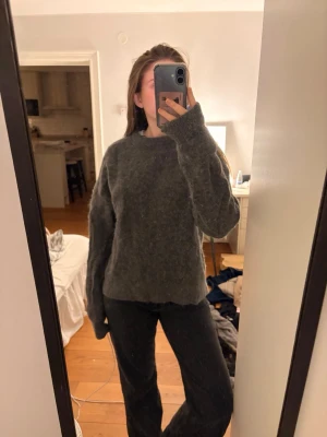 Mörkgrå stickad tröja - Mysig mörkgrå stickad tröja med rund halsringning och långa ärmar. Tröjan har en lite lösare passform och är gjord i ett fluffigt, mjukt material som känns skönt mot huden. Perfekt för kyliga dagar och enkel att matcha med olika outfits.