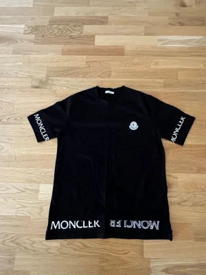 Svart Moncler t-shirt med logga - Snygg svart t-shirt från Moncler med vit logga på bröstet och tryck på båda ärmarna samt nederkanten. Klassisk passform och rund halsringning. Perfekt för dig som vill ha en stilren och trendig look.