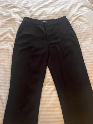 Svarta kostymbyxor från Gina Tricot - Stilrena svarta kostymbyxor från Gina Tricot med raka ben och diskreta sidofickor. Klassisk midja och hällor för skärp. Perfekta för en clean och snygg look. Materialet känns mjukt och har en lätt stretch för extra komfort.