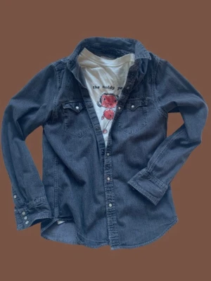 Ralph Lauren Denim&Supply Jeansskjorta - Säljer en vintage grå jeansskjorta med långa ärmar och tryckknappar framtill från Ralph laurens denim&supply. Skjortan har två bröstfickor med lock och knappar. Perfekt att bära öppen över en t-shirt för en cool och avslappnad stil. Mycket bra skick. Skriv gärna för fler bilder/frågor.