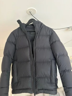 Svart pufferjacka från Peak Performance - Snygg svart pufferjacka från Peak Performance med hög krage och dragkedja framtill. Jackan har en klassisk quiltad design och två sidofickor. Perfekt för kalla vinterdagar och riktigt skön att bära. Materialet är slitstarkt och håller värmen bra.