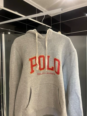 Grå hoodie från Polo Ralph Lauren - Snygg grå hoodie från Polo Ralph Lauren med stor röd POLO-text i mitten. Den är i L men är som M/S eftersom den blivit tvättad fel… Tröjan går att använda vardagligt och passar perfekt ute nu till våren. Vid någon fråga, tveka inte att höra av er!🤝