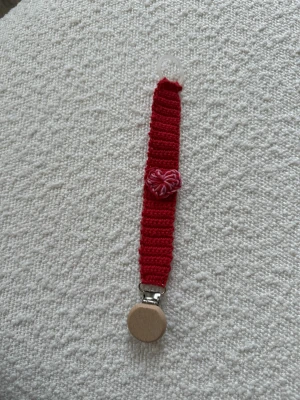 Röd virkad napphållare med hjärta - Handgjord napphållare i röd virkad bomull med ett dekorativt rosa hjärta i mitten. Fästet är i trä och metall, och i andra änden finns en genomskinlig plastring för napp. Perfekt accessoar för att hålla koll på nappen med personlig stil.