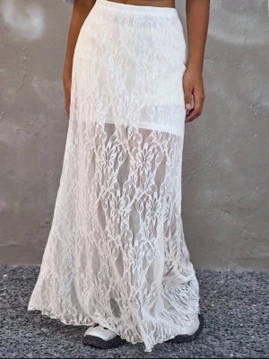 Vit lång spetskjol med blommönster - Säljer en vit långkjol i transparent spets med blommigt mönster. Kjolen har en hög midja och ett tunt innerfoder som slutar ovanför knäna, vilket ger en snygg kontrast till det genombrutna tyget. Perfekt för dig som vill ha en romantisk och trendig look.