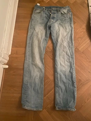 Ljusblå raka jeans från Levi's - Säljer ett par klassiska ljusblå jeans från Levi’s, modell 501, med rak passform och knappgylf. Jeansen är i bra skick med naturligt slitage som ger dem en snygg vintage-look.