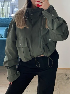 Olivgrön oversized vindjacka - Snygg olivgrön vindjacka med oversized passform och stora fickor framtill. Jackan har hög krage med spänne, dragsko i midjan och tryckknappar på ärmarna. Perfekt för en trendig streetwear-look.