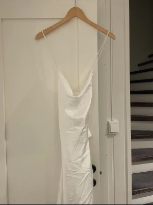 Vit långklänning med tunna band - Elegant vit långklänning med tunna axelband som korsas i ryggen. Klänningen har en draperad urringning och är tillverkad i ett glansigt material som ger en lyxig känsla. Perfekt för dig som vill ha en stilren och trendig look.