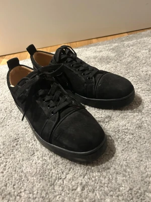 Svarta skor från Louboutin - Säljer ett par svarta sneakers från Christian Louboutin i mocka med klassisk röd sula och rund tå. Skorna har snörning och diskret design som ändå sticker ut tack vare den ikoniska röda undersidan. Insidan är i ljust skinn för extra komfort. Påse och skohornet ingår obs sömnen i öppningen är lite öppet men går säker o fixa!