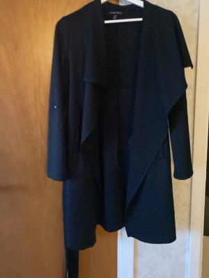 Svart kappa  - Svart kappa från AMISU. Kappan har långa ärmar, två fickor och ett bälte i midjan. Använd några gånger men i bra skick. Passar bra till vår och höst.