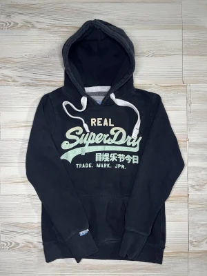 Svart Superdry hoodie med tryck - Svart hoodie från Superdry med stor logga och text i grönt och vitt på bröstet🤩Hoodien är i fint skick och passar Xs-S🙌🏻 Hör av er vid funderingar samt snabbaffär😇