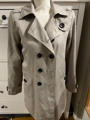Beige trenchcoat med dubbelknäppning - Stilren beige trenchcoat med klassisk dubbelknäppning och stora svarta knappar. Jackan har bred krage, axelklaffar och justerbara band vid ärmsluten. Tillverkad i ett slätt tyg som ger en snygg och tidlös look. Perfekt för dig som vill ha en ikonisk ytterjacka. Finns tyvärr inget bälte till