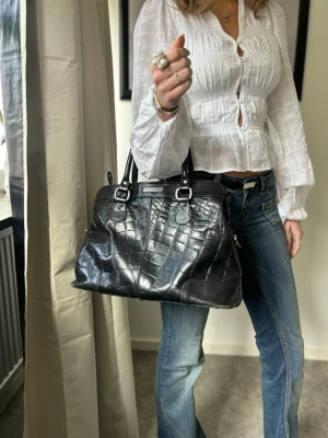 Svart handväska i crocopräglat läder - Snygg svart handväska i crocopräglat läder med dubbla handtag och silvriga metalldetaljer. Väskan har en klassisk, rymlig form och stängs med dragkedja. Perfekt för dig som gillar en stilren och trendig accessoar. Finns några små slitningar på undersidan.