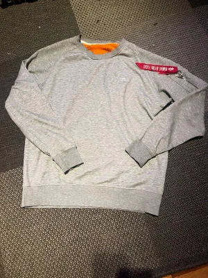 Grå sweatshirt från Alpha Industries - Snygg grå sweatshirt från Alpha Industries med rund halsringning och ribbade muddar. Tröjan har en ikonisk röd tagg med texten 'Remove Before Flight' på ärmen och en liten ficka med dragkedja. Insidan har orange detalj. Perfekt för en casual look.