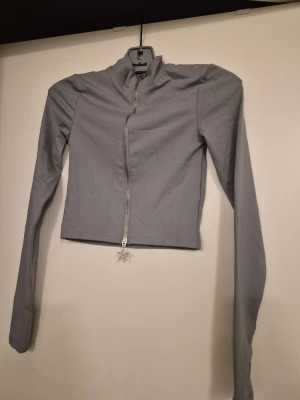 Ljusgrå ziptröja med stjärndragkedja - Snygg ljusgrå ziptröja med hög krage och långa ärmar. Dragkedjan går snett över framsidan och har en unik stjärnformad dragkedjehänge. Tröjan är figurnära och croppad, perfekt till jeans eller kjol. Materialet känns mjukt och stretchigt.