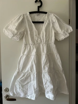 Vit kort klänning med puffärm - Söt vit kortklänning med v-ringning och puffärmar. Klänningen har volangdetaljer och är tillverkad i bomull, vilket gör den luftig och bekväm. Perfekt för varma dagar och enkel att styla med accessoarer.