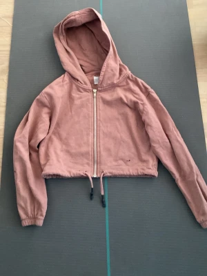 Rosa cropped hoodie med dragkedja - Säljer en rosa cropped hoodie med dragkedja framtill och justerbar dragsko i midjan. Hoodien har långa ärmar, huva och ribbade muddar vid ärmsluten. Perfekt för en avslappnad och trendig look.