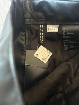 Svart kort kjol i skinnimitation H&M - Säljer en svart kort kjol från H&M Divided i skinnimitation. Kjolen har en dragkedja bak och volangdetalj längst ner. Perfekt för dig som vill ha en edgy och trendig look.