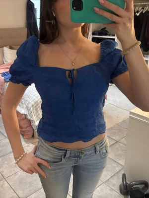 Blå croppad blus med puffärmar - Superfin blå croppad blus med puffärmar och söt knytning framtill. Fyrkantig urringning och lätt vågig nederkant ger en trendig look. Perfekt att styla med jeans eller kjol för en cool vibe.