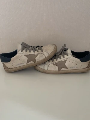 Golden Goose vita sneakers med stjärna - Golden Goose sneakers i vitt läder med grå stjärna på sidan, grå skosnören och mörkblå detalj vid hälen. Klassisk låg modell med slitna detaljer och GGDB/SSTAR-tryck på sidan. Snygg retrostil som sticker ut.