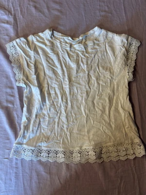Vit topp med spetsdetaljer - Söt vit topp med spets längs ärmslut och nederkant. T-shirttyp med korta ärmar och rund halsringning. Perfekt för dig som gillar romantiska detaljer och enkel stil. Materialet känns mjukt och luftigt, troligen bomull.