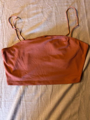 Rostfärgad ribbad linnetopp - Trendig rosafärgad linnetopp med tunna axelband och ribbad struktur. Toppen är croppad och har en enkel, stilren design som passar perfekt till höga jeans eller kjol. Materialet känns mjukt och följsamt mot huden.