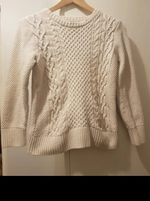 Beige kabelstickad tröja - Snygg beige stickad tröja med kabelstickat mönster och strukturstickade partier. Tröjan har rund halsringning, långa ärmar och breda ribbade muddar vid ärmslut och nederkant. Perfekt för lager på lager och en klassisk look. Storlek:170