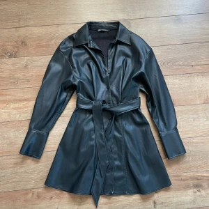 Skinn Skjorta  - Snygg svart skjorta /  klänning från Zara i skinnimitation med knappar framtill och bälte i midjan. Klänningen har långa ärmar med breda manschetter och klassisk krage. Perfekt för dig som gillar en edgy och trendig look.