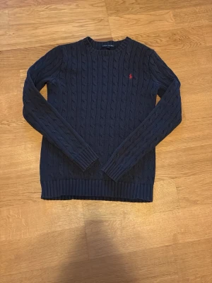 Mörkblå kabelstickad tröja Ralph Lauren - Kabelstickad mörkblå tröja från Ralph Lauren med rund halsringning och den klassiska röda logotypen broderad på bröstet. Tröjan har långa armar och ribbade muddar vid ärmslut och nederkant. Perfekt för en stilren och tidlös look. Jag säljer den eftersom den börjar bli för liten speciellt i armarna. Den är i fint skick och perfekt nu med en väst på. Pris kan diskuteras.