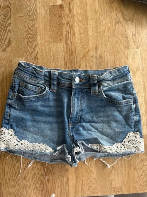 Blåa Jeansshorts med detaljer  - Supersnygga jeansshorts i blå denim med slitna detaljer och fin spets längs kanten som ger en feminin och trendig look ✨! VITA FLÄCKAR VID BAKFICKA!  ✔️ Storlek 170 men passar mig som bär XS/S ✔️ Fint skick ✔️ Råa, fransiga benslut för en cool stil ✔️ Dekorativ spets som gör dem extra speciella  Perfekta till sommaren – lika snygga med ett enkelt linne som en söt topp 🌞  📦 Snabb leverans 💬 Hör av dig vid frågor eller fler bilder 