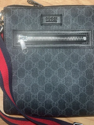 Svart axelväska från Gucci - Snygg svart axelväska från Gucci med klassiskt GG-mönster och silverfärgad dragkedja framtill. Väskan har en justerbar axelrem i rött och marinblått tyg. Perfekt för att bära det viktigaste med stil.