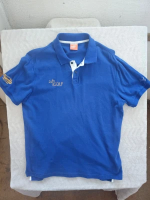 Blå pikétröja från Puma - Blå pikétröja från Puma med vit knappslå och broderad text '24h GOLF' i guld på bröstet. Klassisk krage och korta ärmar. Perfekt för dig som gillar sportig stil och vill ha något bekvämt och snyggt.