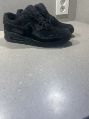 Svarta Nike Air Max 90 sneakers - Snygga helsvarta Nike Air Max 90 sneakers med klassisk siluett. Skorna har en mix av läder och mesh, ikonisk Air-dämpning i sulan och diskreta Nike-loggor på sidan och hälen. Perfekta för dig som gillar en clean och sportig look.