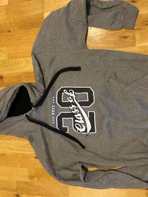 Grå hoodie Class of 22 tryck stl L bra skick - Snygg grå hoodie med svart och vitt tryck 'Class of 22' på bröstet. Hoodien har svarta dragsnören, känguruficka och mjukt material. Perfekt för dig som gillar college-stil och vill ha en bekväm och stilren tröja.